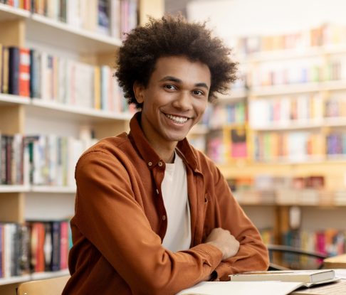 A smiling young man sitting at a table, surrounded by bookshelves filled with books.